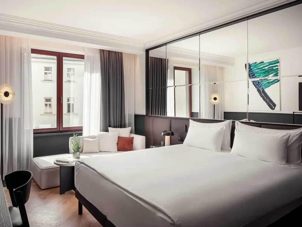 Modernes Hotelzimmer mit großem Bett, verspiegelter Wand, neutralem Dekor und abstraktem Kunstwerk - dieses Almanac verfügt über einen gemütlichen Sitzbereich am Fenster und ein privates Zimmer mit Balkon für einen entspannenden Ausblick.