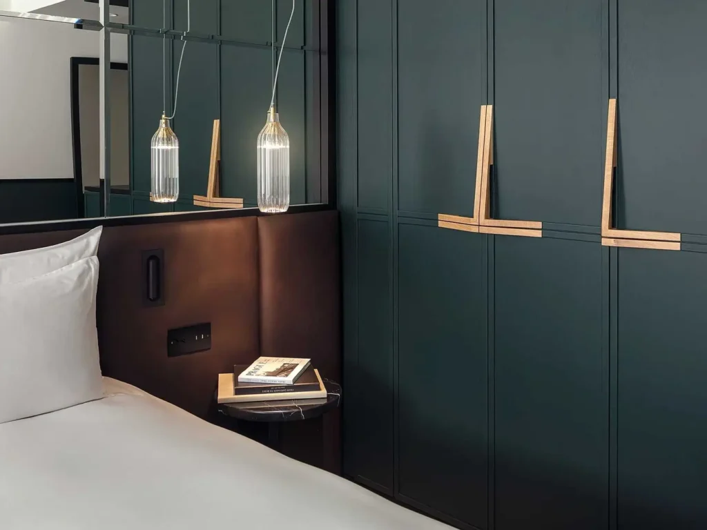 Ein modernes Deluxe-Zimmer mit einem weißen Bett, einem kleinen Beistelltisch mit Büchern, Hängelampen und dunkelgrün getäfelten Wänden mit eleganten Holzgriffen.