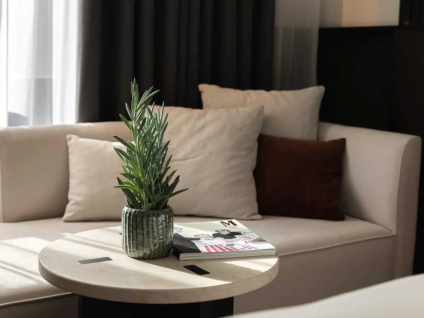 Eine kleine Topfpflanze und Zeitschriften stehen auf einem runden Tisch vor einem beigen Sofa mit weißen und braunen Kissen und bilden eine gemütliche Ecke, die an ein Deluxe Zimmer erinnert.