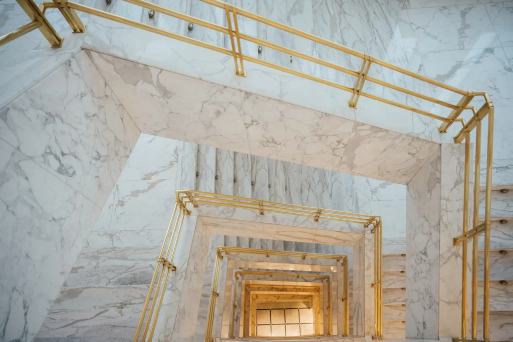 Eine Marmortreppe mit goldenen Handläufen bildet eine geometrische Spirale, die in einem modernen, hell erleuchteten Innenraum von unten betrachtet wird - perfekt, um besondere Angebote zu präsentieren.