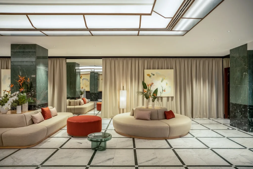 Moderne Hotellobby mit Marmorböden, grünen Marmorsäulen, beigen Sofas, orangefarbenen Kissen und Hockern, Glastisch, großen Kunstwerken und neutralen Vorhängen - perfekt für Kulturveranstaltungen oder zum stilvollen Entspannen.