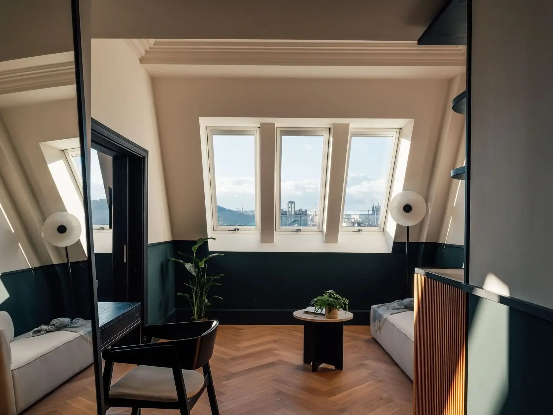 Experience the Almanac’s Castle View Suite: a modern room with three tall windows, light wood herringbone floor, a black chair, two sofas, and a round coffee table with plants—plus stunning cityscape views outside.