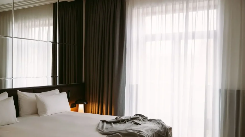Ein ordentlich gemachtes Bett mit weißer Bettwäsche und Kissen, eine graue Decke am Fußende und große Fenster mit durchsichtigen und dunklen Vorhängen, die natürliches Licht hereinlassen, schaffen ein ruhiges Ambiente in der Executive Suite.