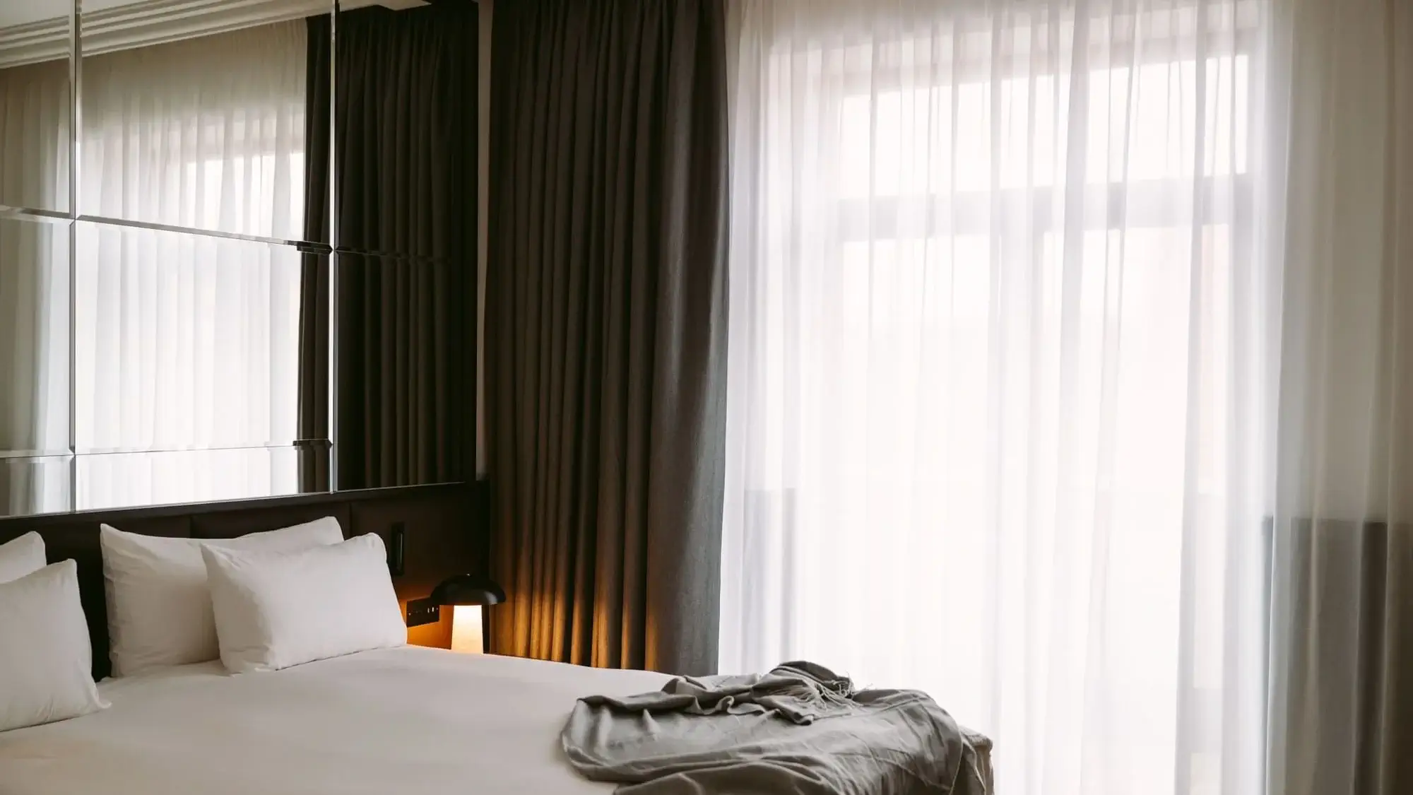 Ein ordentlich gemachtes Bett mit weißer Bettwäsche und Kissen, eine graue Decke am Fußende und große Fenster mit durchsichtigen und dunklen Vorhängen, die natürliches Licht hereinlassen, schaffen ein ruhiges Ambiente in der Executive Suite.