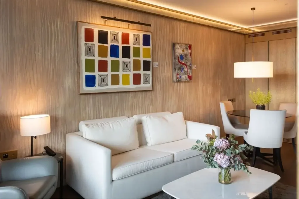 Moderno salón con un sofá blanco, arte abstracto en la pared, una mesa de centro con flores y una zona de comedor con una mesa redonda y una lámpara colgante al fondo.