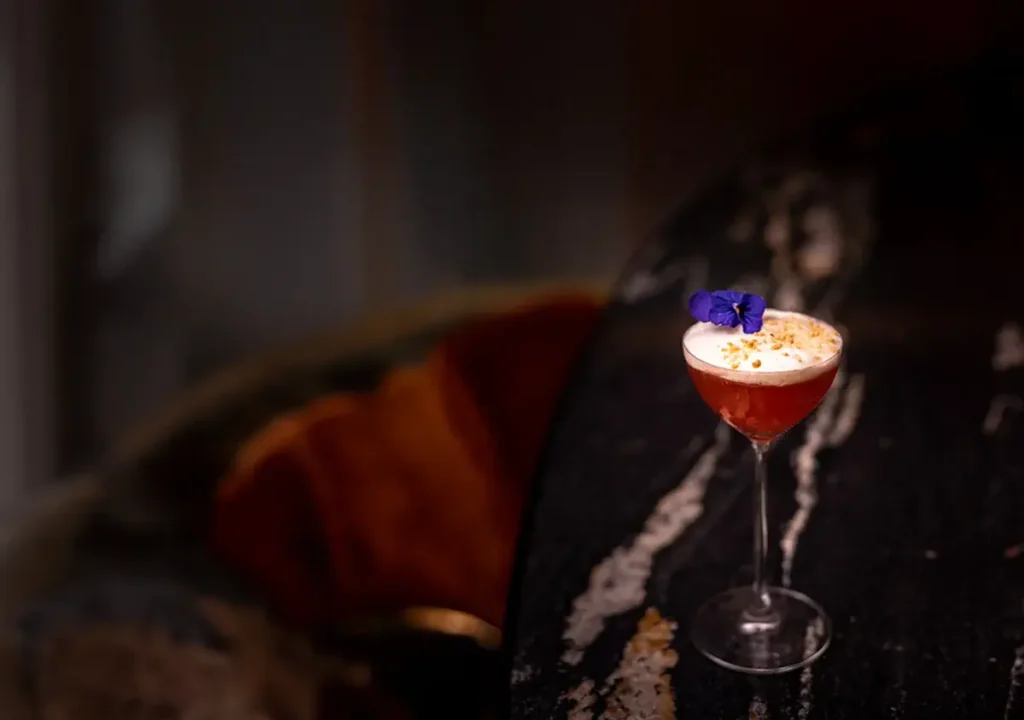 Ein Cocktail in einem Coupe-Glas mit Schaumkrone, garniert mit einer violetten Blume, steht auf einer dunklen Marmorplatte - eine kunstvolle Mischung aus Getränke- und Speisenästhetik.