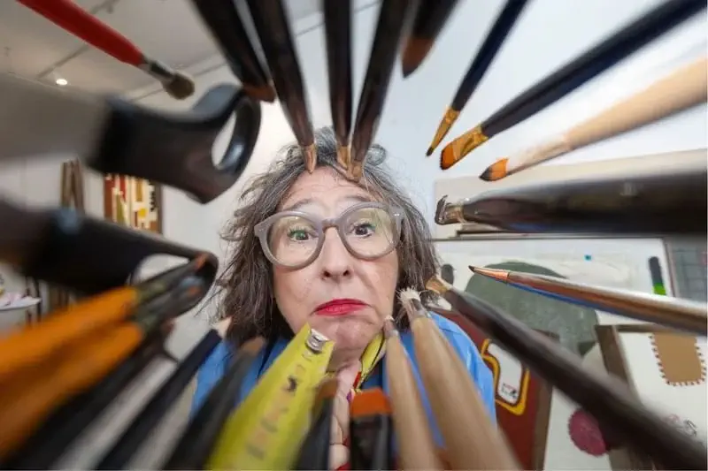 Una persona amb els cabells grisos i ulleres mira a través d'un grup de pinzells i llapis d'art, amb la cara emmarcada per les eines en primer pla.