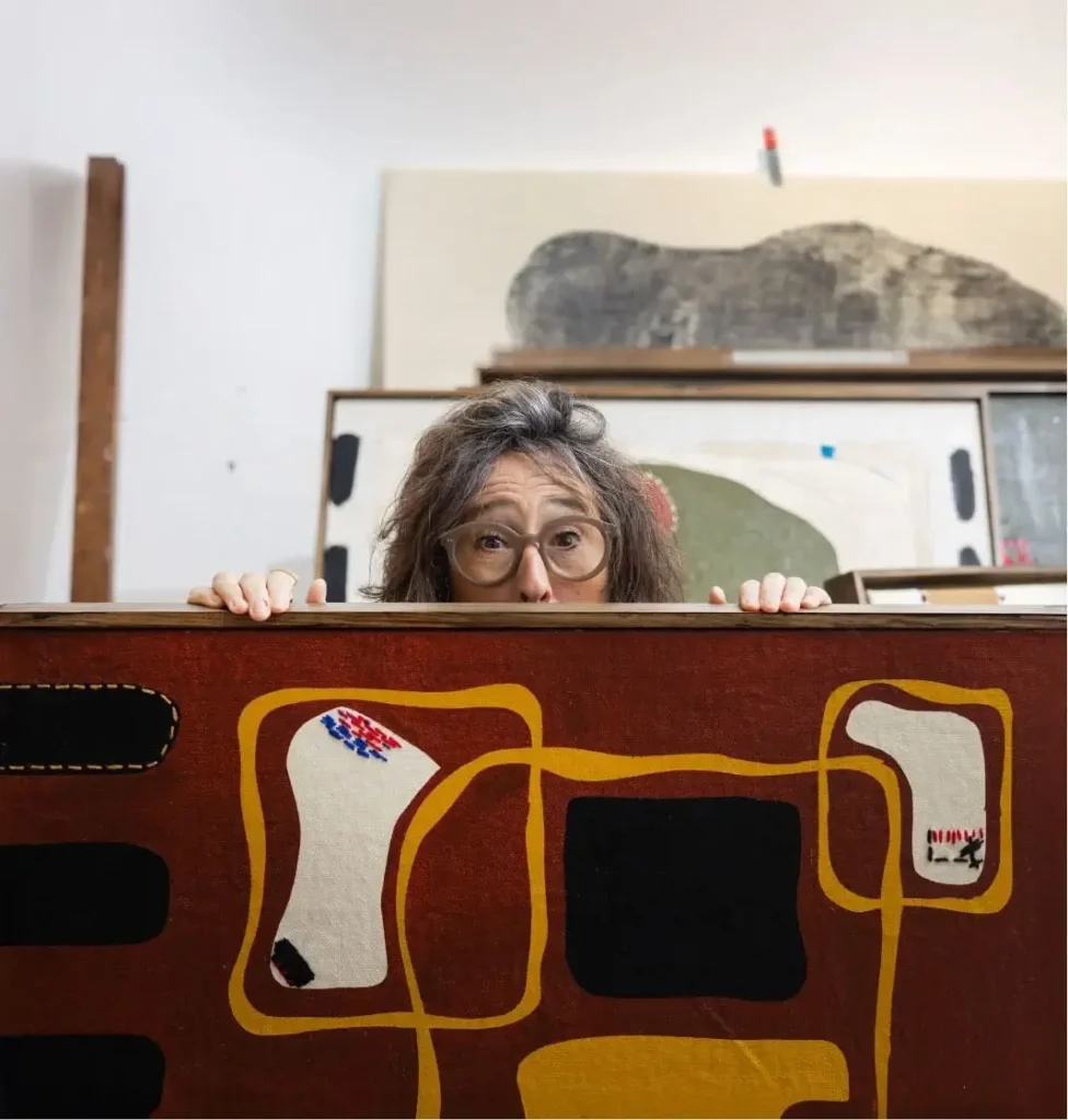 Una persona con gafas mira por encima de un colorido cuadro abstracto en un estudio de arte con lienzos al fondo.