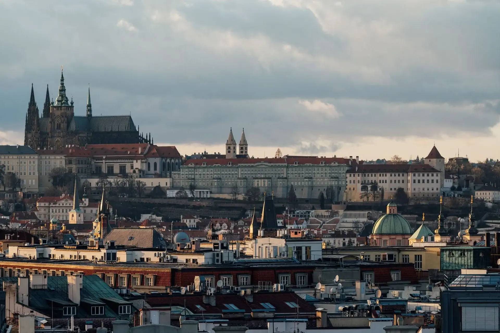 Stadtansicht von Prag mit historischen Gebäuden, roten Dächern, Kirchtürmen und der Prager Burg, die unter einem wolkenverhangenen Himmel zu sehen ist - Heimat charmanter Cafés und einzigartiger Concept Stores inmitten der historischen Straßen.