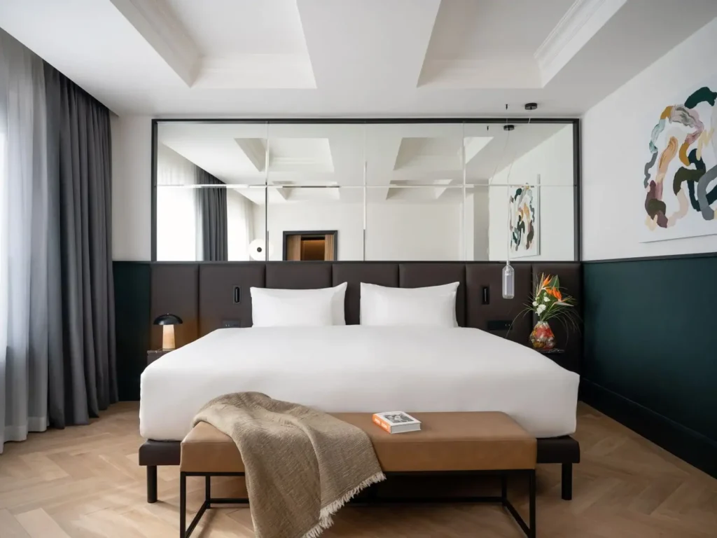Eine moderne Präsidentensuite mit einem großen Bett mit weißer Bettwäsche, einer braunen Bank mit einer beigen Decke, einem Nachttisch mit Lampe und abstrakten Kunstwerken an den Wänden.
