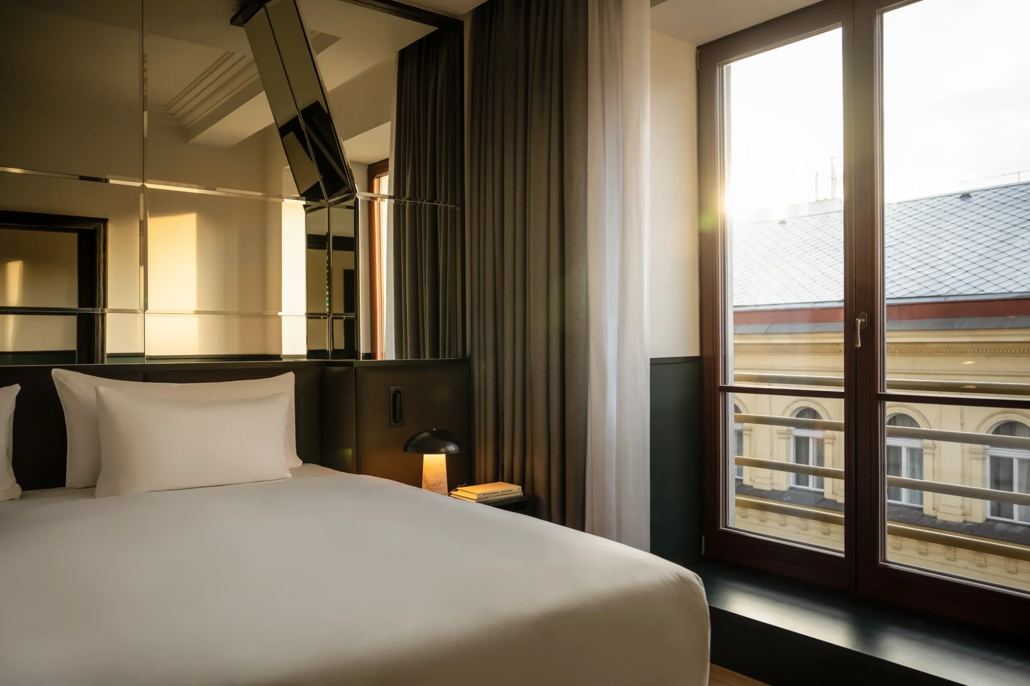 Moderne Executive Suite mit sauber gemachtem Bett, verspiegelter Wand, Nachttischlampe und großem Fenster mit Blick auf ein Gebäude bei Sonnenuntergang.