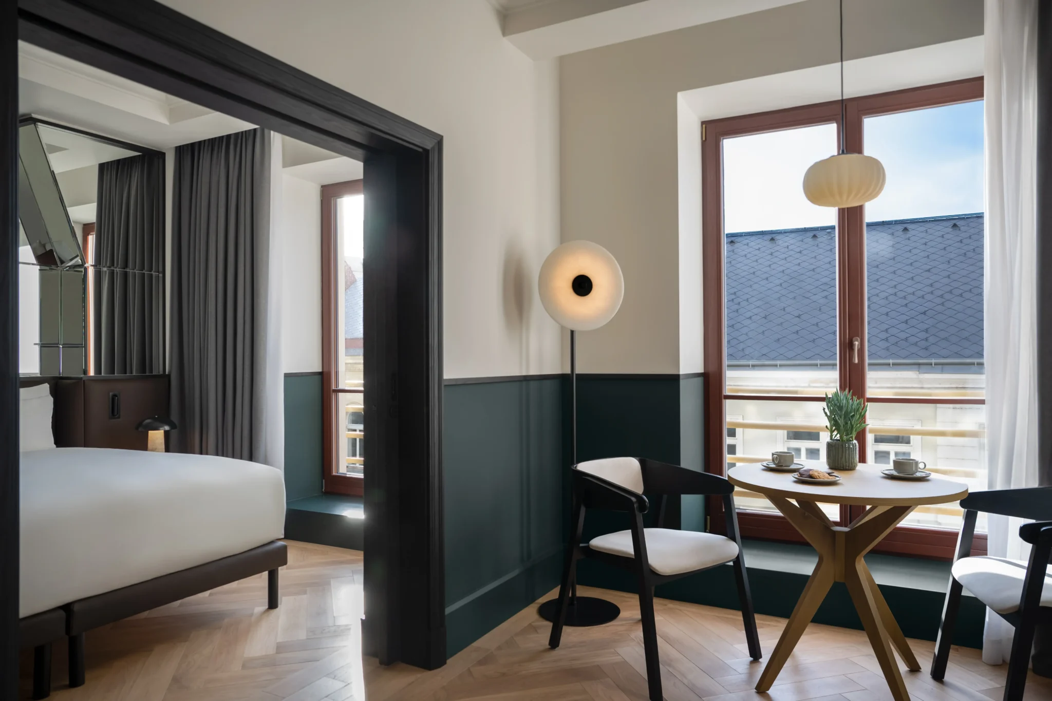Moderne Executive Suite mit einem Bett, einem runden Tisch, zwei Stühlen und großen Fenstern, die den Raum mit natürlichem Licht erfüllen. Kaffeetassen und eine Blumenvase stehen elegant auf dem Tisch.