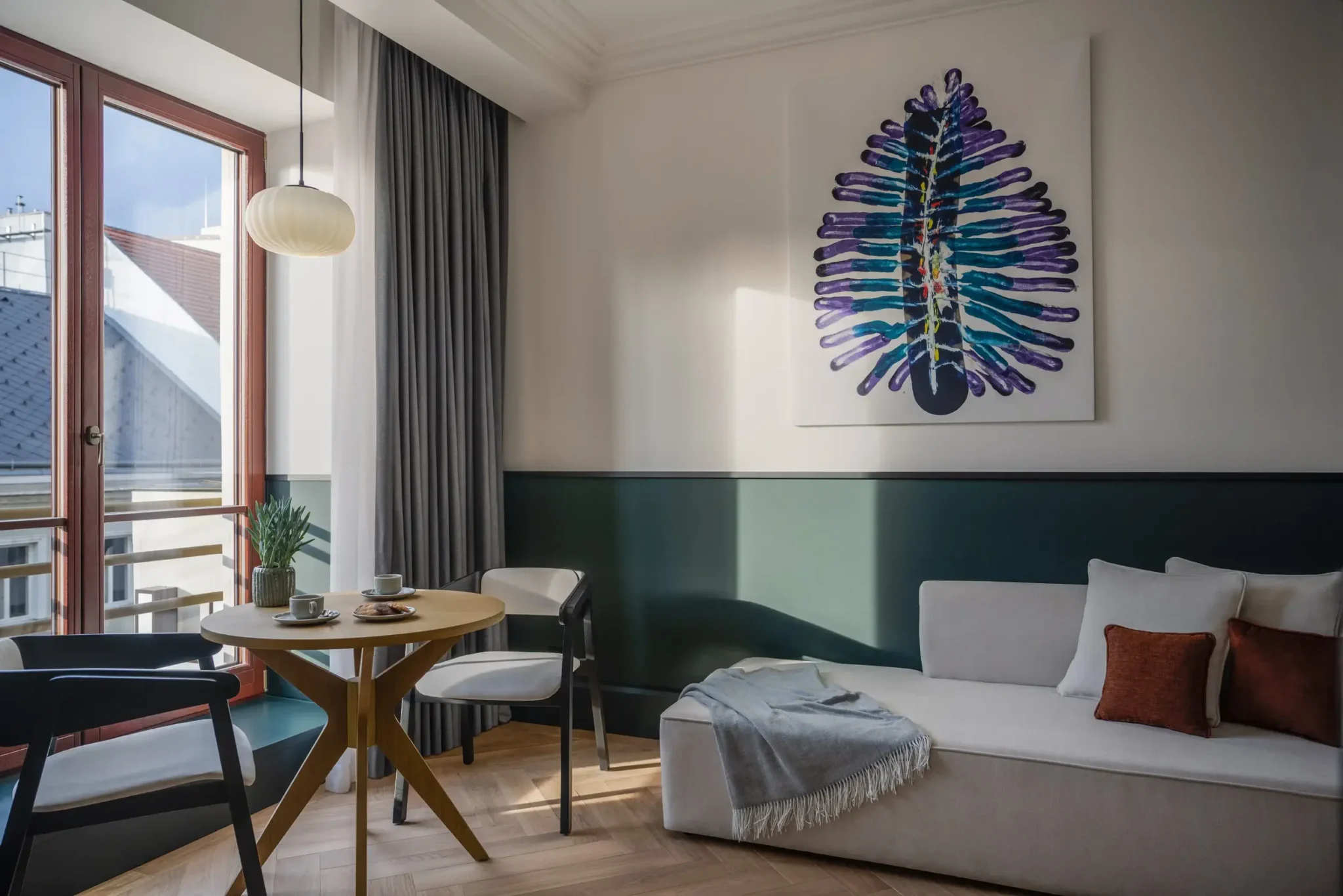 Modernes Wohnzimmer der Executive Suite mit rundem Tisch, Stühlen, einem hellen Sofa mit Kissen und einer Decke, einem großen Fenster und einem abstrakten Kunstwerk an der Wand.