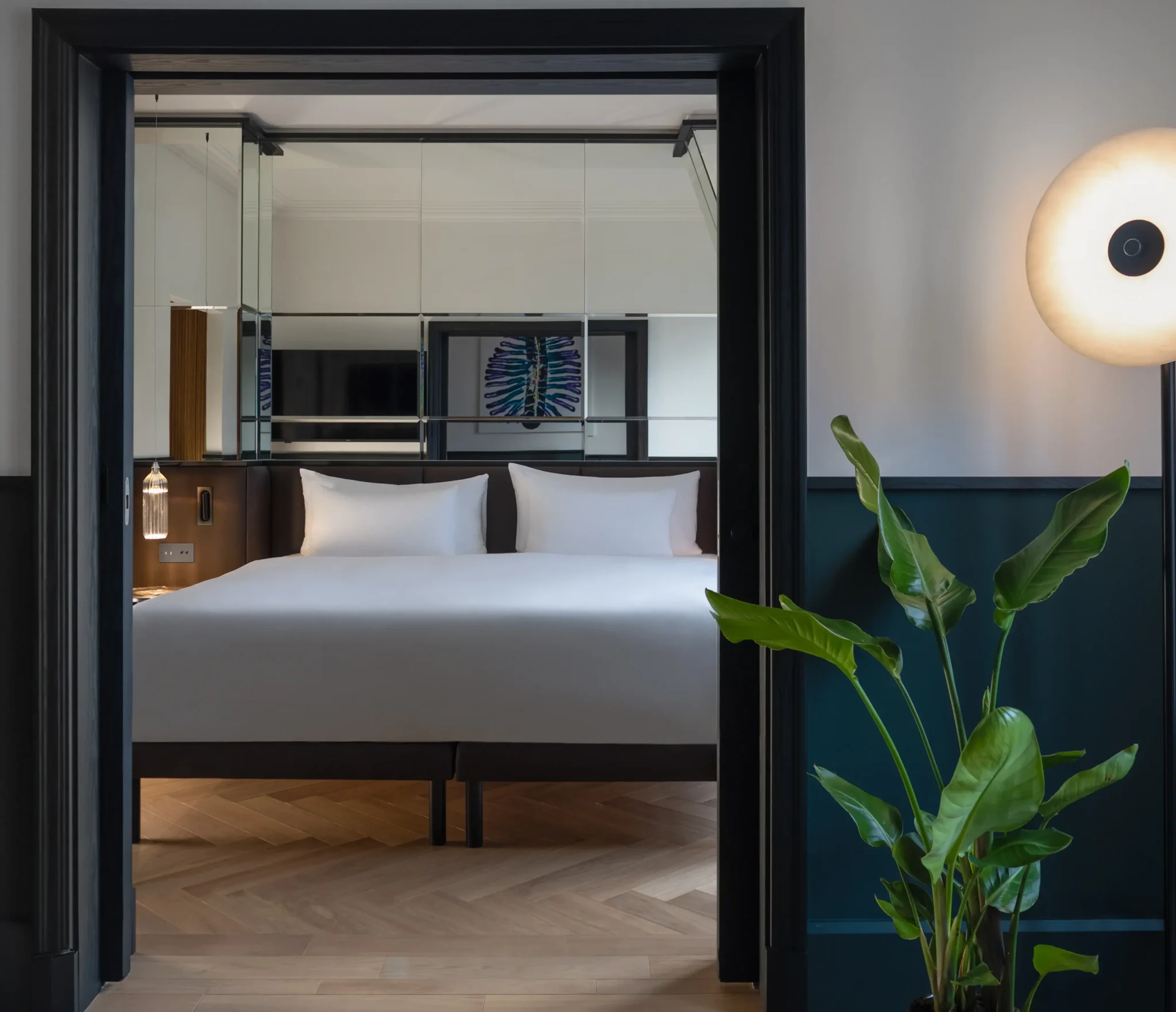 Eine Executive Suite mit einem großen Bett mit weißem Bettzeug, einer Spiegelwand, einem Holzboden, einer Topfpflanze und einer runden Stehlampe schafft eine moderne und einladende Atmosphäre.