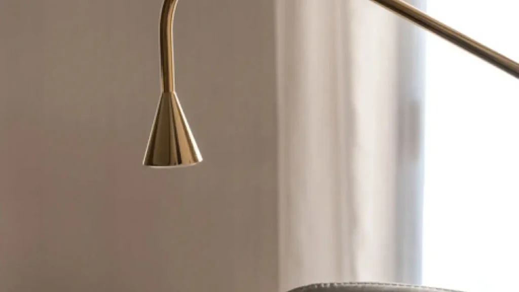 Close-up of a metallic, curved lamp with a cone-shaped shade beside a white curtain and part of a gray chair.