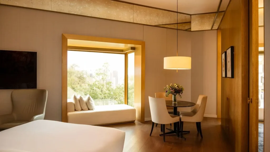 Erleben Sie die Almanac Family Suite: ein modernes Hotelzimmer mit einem Bett, einem Fensterplatz, einem runden Tisch mit vier Stühlen, einer Pendelleuchte und großen Fenstern mit Blick ins Grüne.