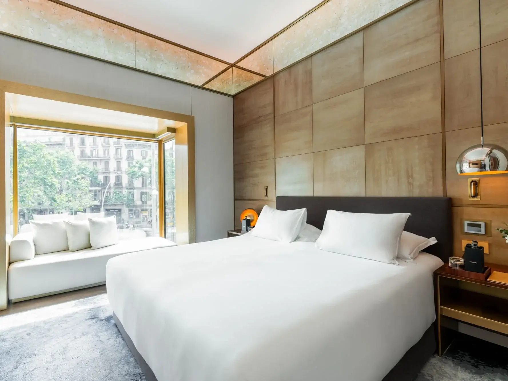 Moderne Familiensuite mit einem großen Bett, zwei Nachttischen, Hängelampen und einem Fensterplatz mit Blick auf eine Stadtlandschaft mit Bäumen.