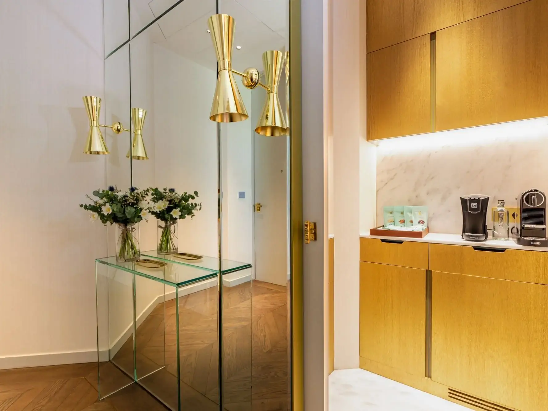 Ein moderner Flur in der Familiensuite ist mit einem gläsernen Konsolentisch, Blumenarrangements, Wandleuchten aus Messing und einer angrenzenden Küchenzeile mit goldenen Schränken und Kaffeemaschinen ausgestattet.