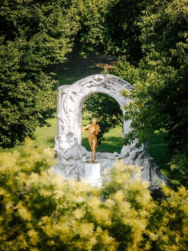 Eine goldene Statue von Johann Strauss, die seinen 200. Geburtstag feiert, steht in einem weißen, verschnörkelten Steinbogen, umgeben von üppig grünen Bäumen und Blättern.