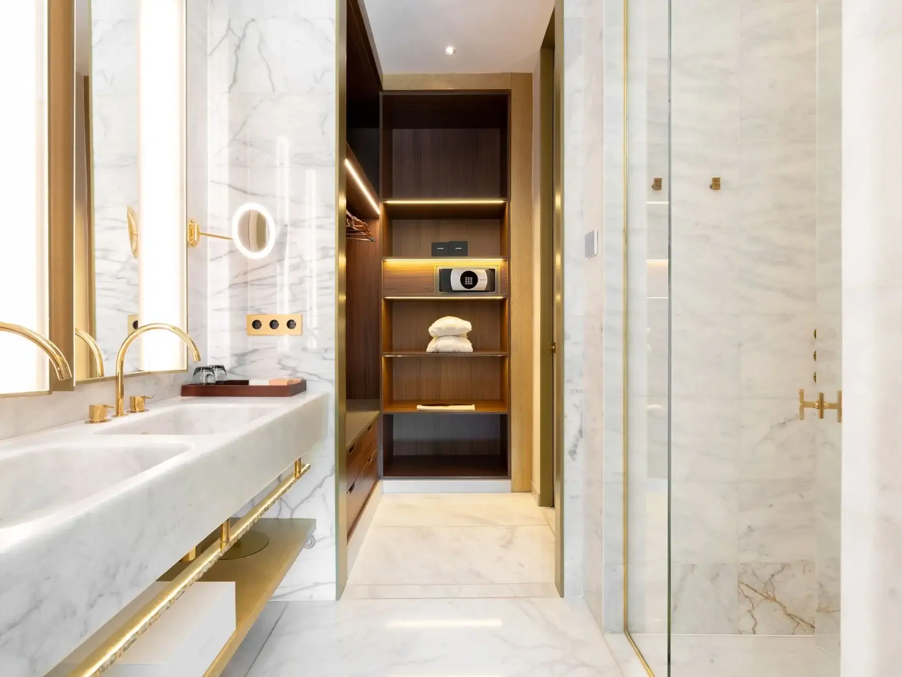 Modernes Badezimmer in der Almanac Family Suite mit Marmorwänden und -böden, Doppelwaschbecken mit goldenen Armaturen, Glasdusche sowie einem offenen Schrank mit Regalen, Handtüchern und einem Safe.