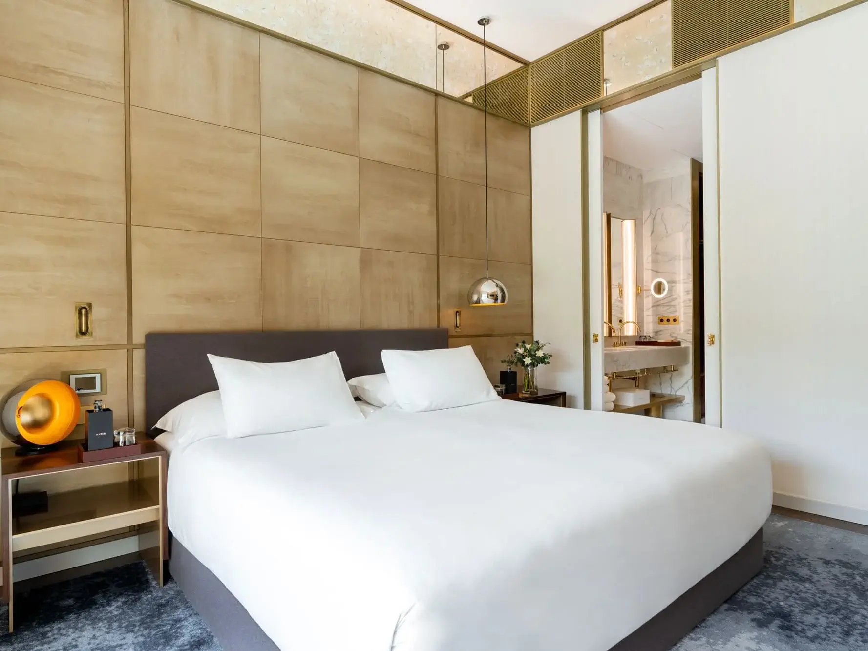 Modernes Hotelzimmer mit großem Bett, weißer Bettwäsche, holzgetäfelten Wänden, Nachttischen, modernen Lampen und einem offenen Durchgang zum Badezimmer - perfekt zum Entspannen in der einladenden Almanac Family Suite.