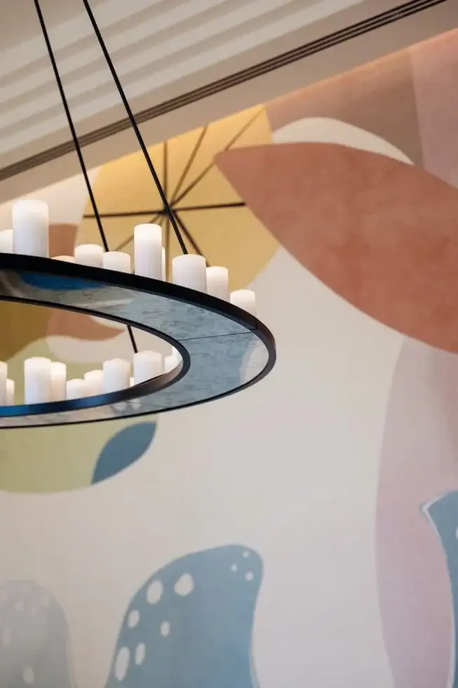 A modern circular chandelier with unlit white candles hangs in front of a pastel abstract mural featuring organic shapes and soft colors.