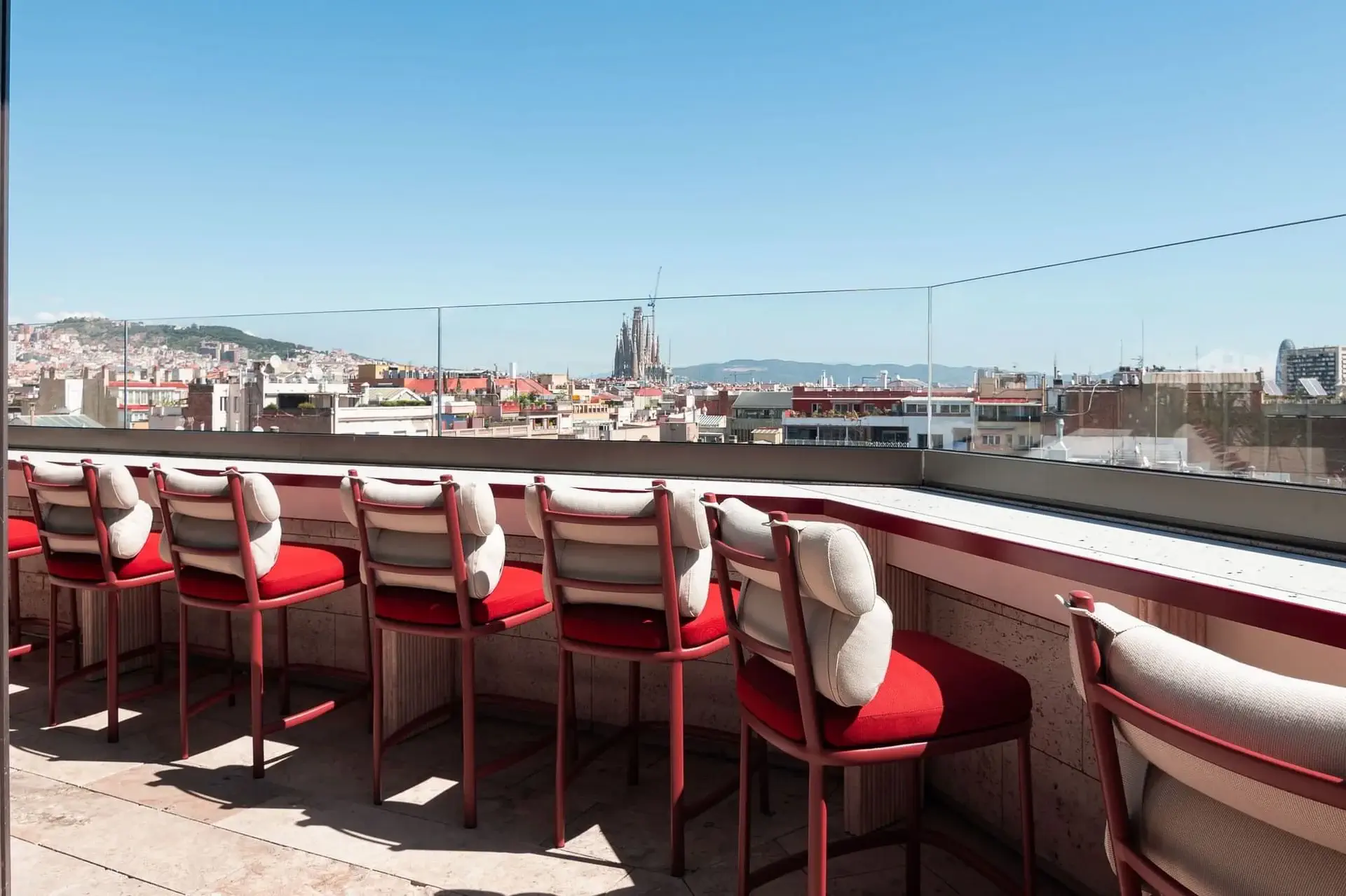 Una terrassa al terrat de Terrassa Azimuth compta amb cadires vermelles amb coixins i vistes a un paisatge urbà sota un cel blau clar, amb una catedral notable visible en la distància.