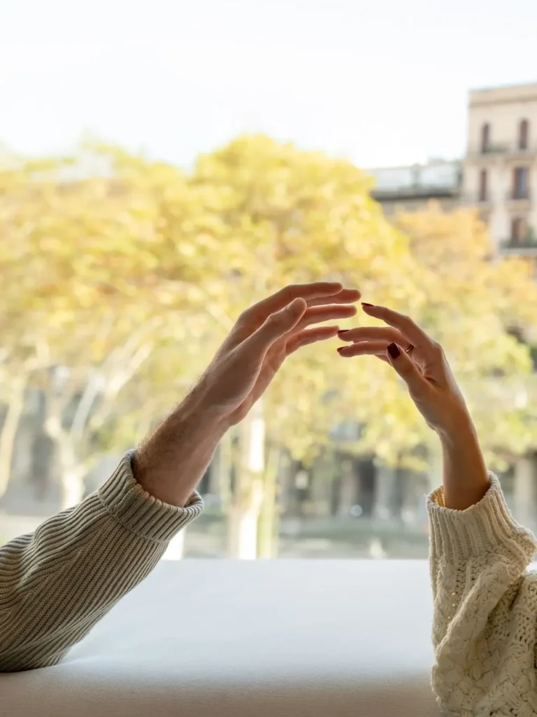 Two people in sweaters reach toward each other with open hands, set against a blurred outdoor background with autumn trees and distant buildings.