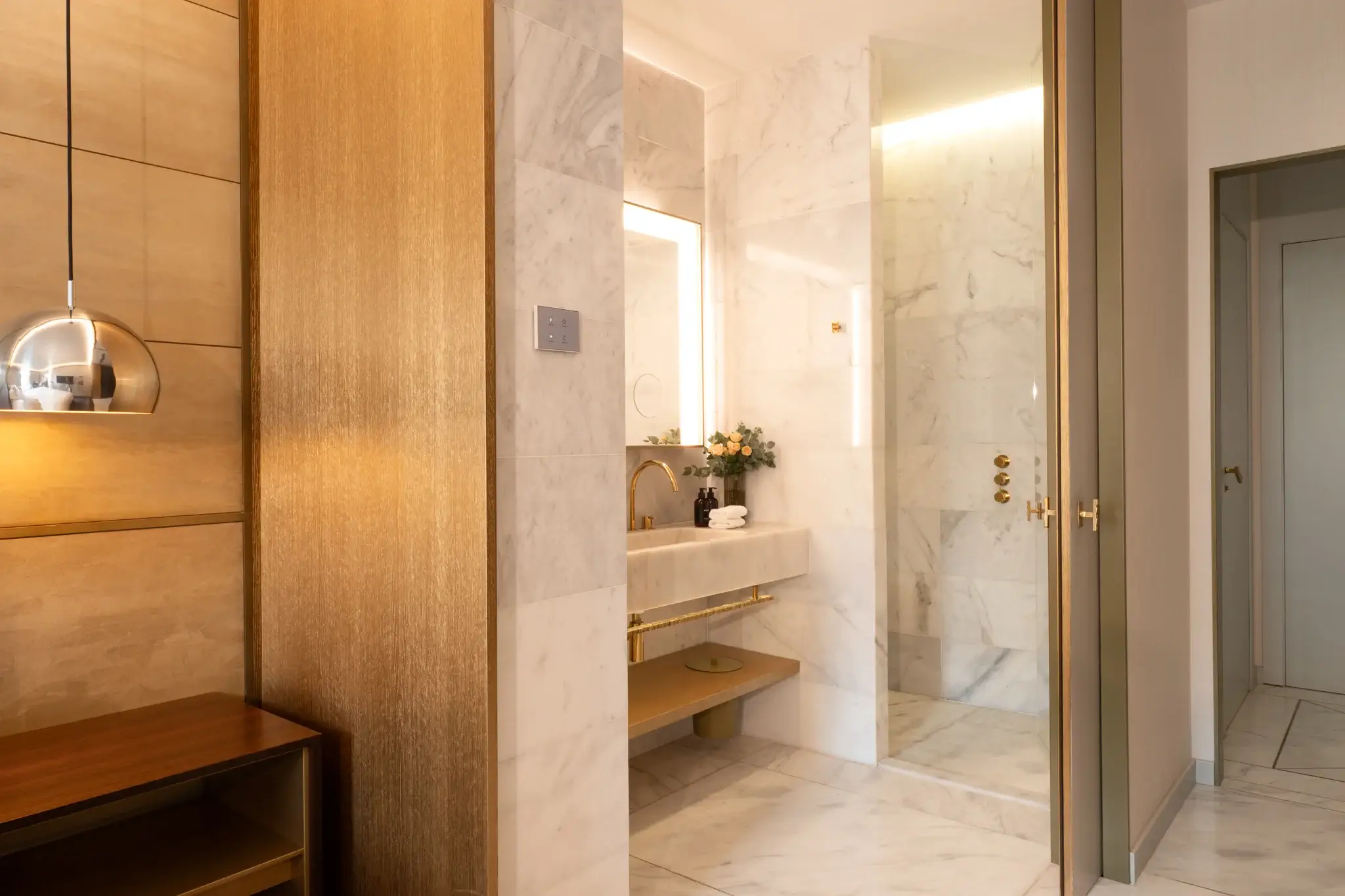 Modernes Badezimmer mit Marmorwänden und -boden, goldenen Armaturen, einem Spiegel über dem Waschbecken und einer Glasduschkabine. Auf der linken Seite sind warme Beleuchtung und Holzakzente zu sehen.