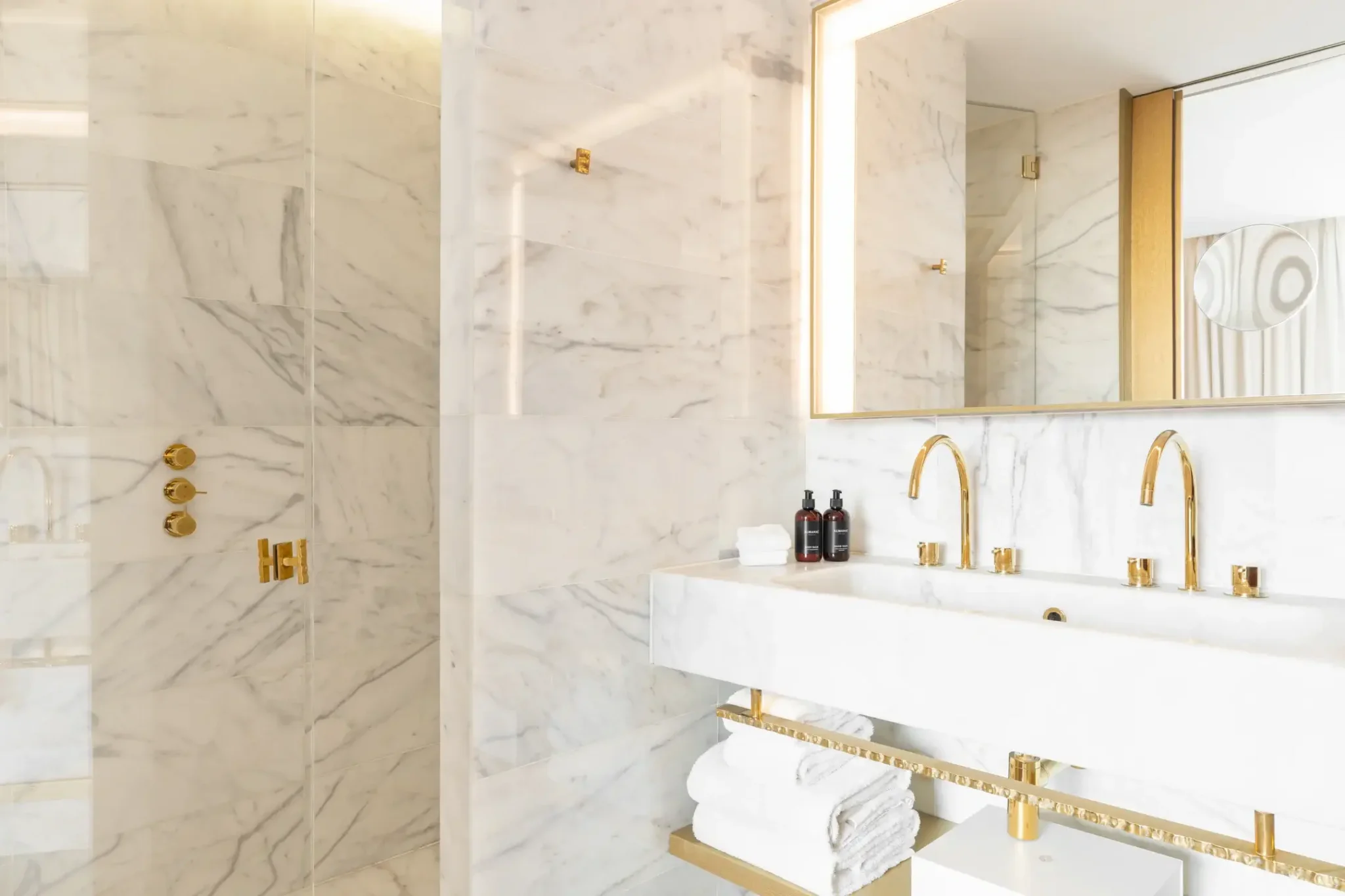 Modernes Badezimmer mit weißen Marmorwänden, goldenen Armaturen, einem Doppelwaschbecken, einem Spiegel, Handtüchern auf einem Regal und Toilettenartikeln auf dem Tresen.