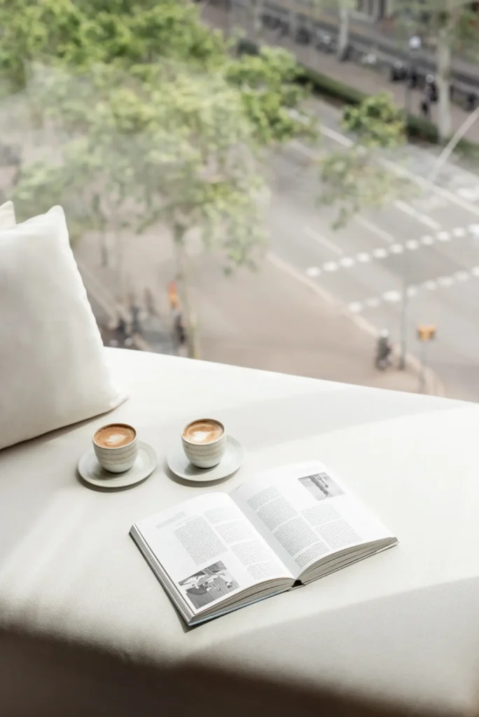 Dues tasses de cafè i un llibre obert descansen sobre una taula blanca al costat d'una finestra, convidant-vos a gaudir d'una Escapada de Tardor mentre observeu els carrers de la ciutat plens d'arbres i trànsit.