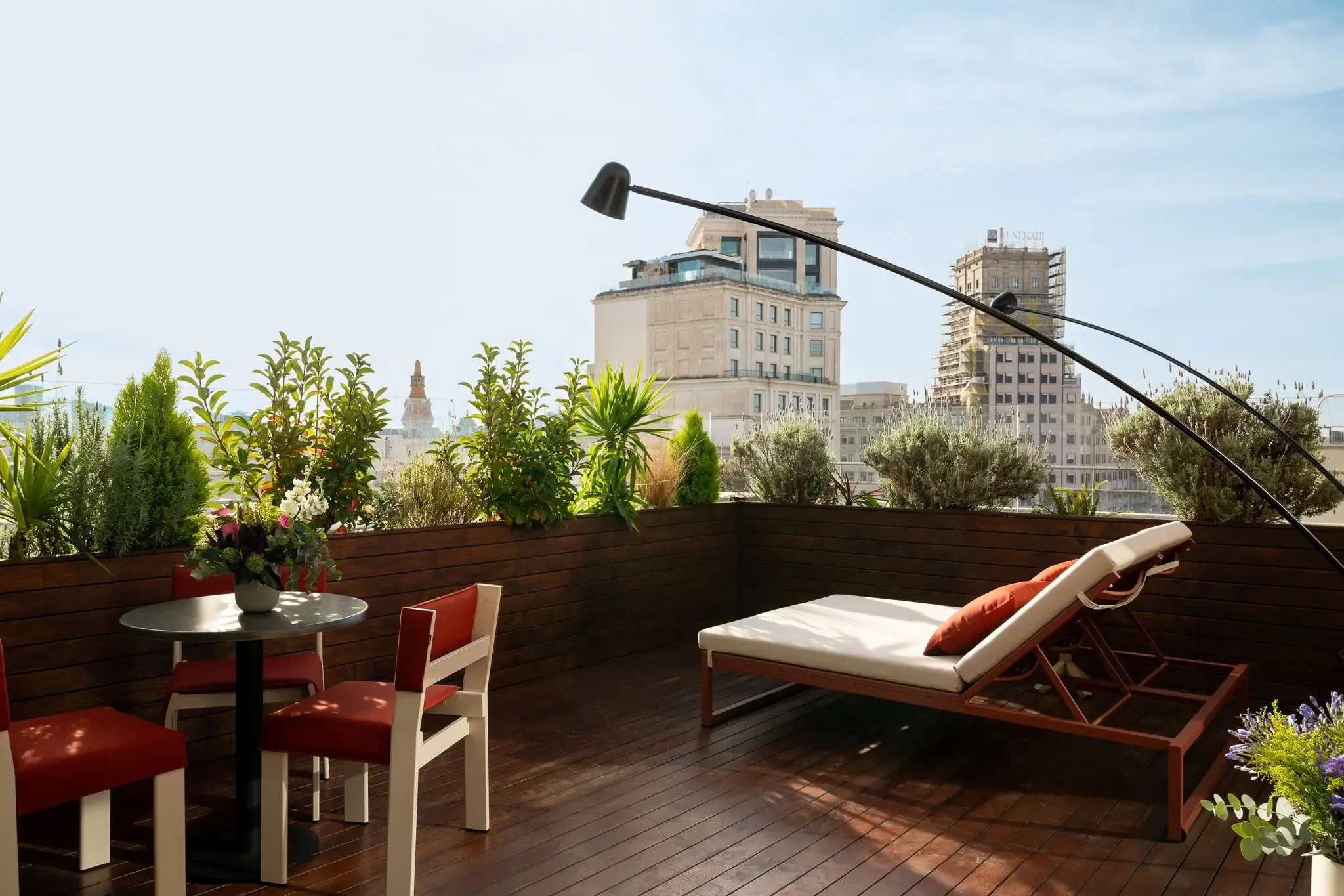 Die Dachterrasse dieser Luxussuite verfügt über einen Holzboden, einen Loungesessel, einen kleinen Tisch mit zwei Stühlen, Topfpflanzen und die Gebäude der Stadt im Hintergrund unter einem klaren Himmel.
