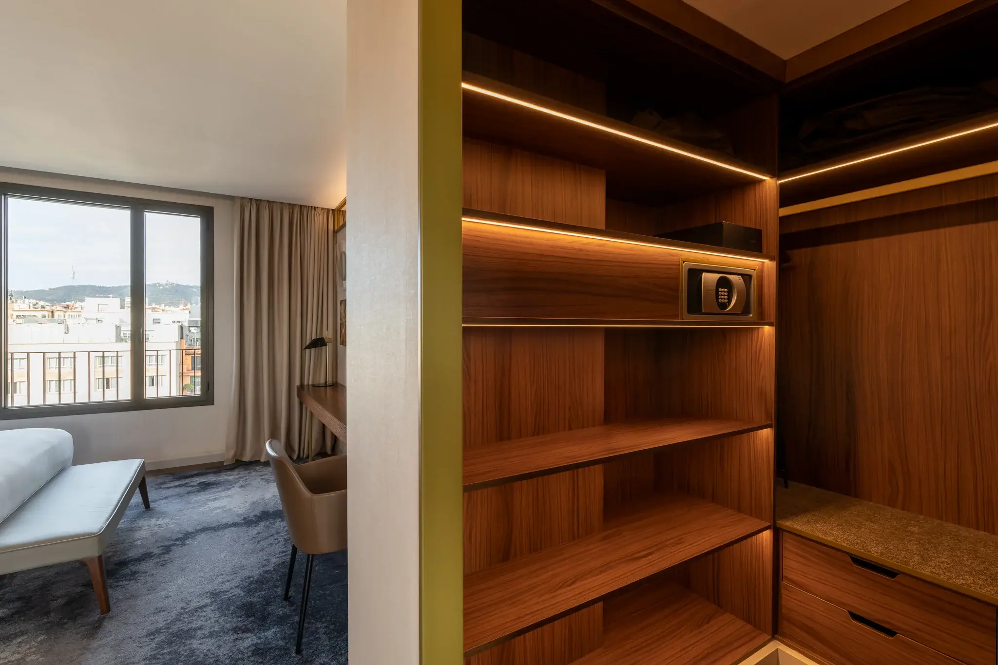 Eine moderne Penthouse-Suite mit herrlichem Blick auf die Stadt, ausgestattet mit einem Bett, einem Schreibtisch und einem offenen Holzschrank mit Regalen und einem Safe.