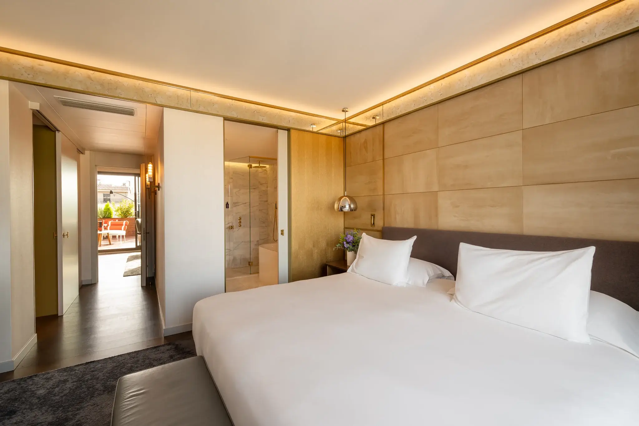 Eine moderne Penthouse-Suite mit einem großen Bett, neutralen Farbtönen, Glasschiebetüren zum Badezimmer und einer offenen Tür, die zu einer Terrasse mit Sitzgelegenheiten im Freien führt.
