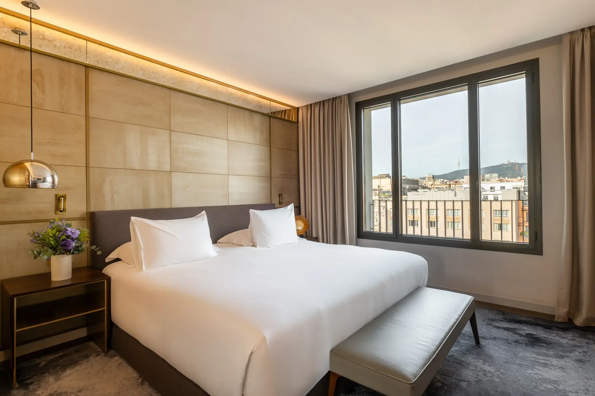 Eine moderne Penthouse-Suite mit einem großen Bett, zwei Nachttischen, einer Bank am Fußende des Bettes und einem Fenster, das einen Blick auf die Stadt bietet.