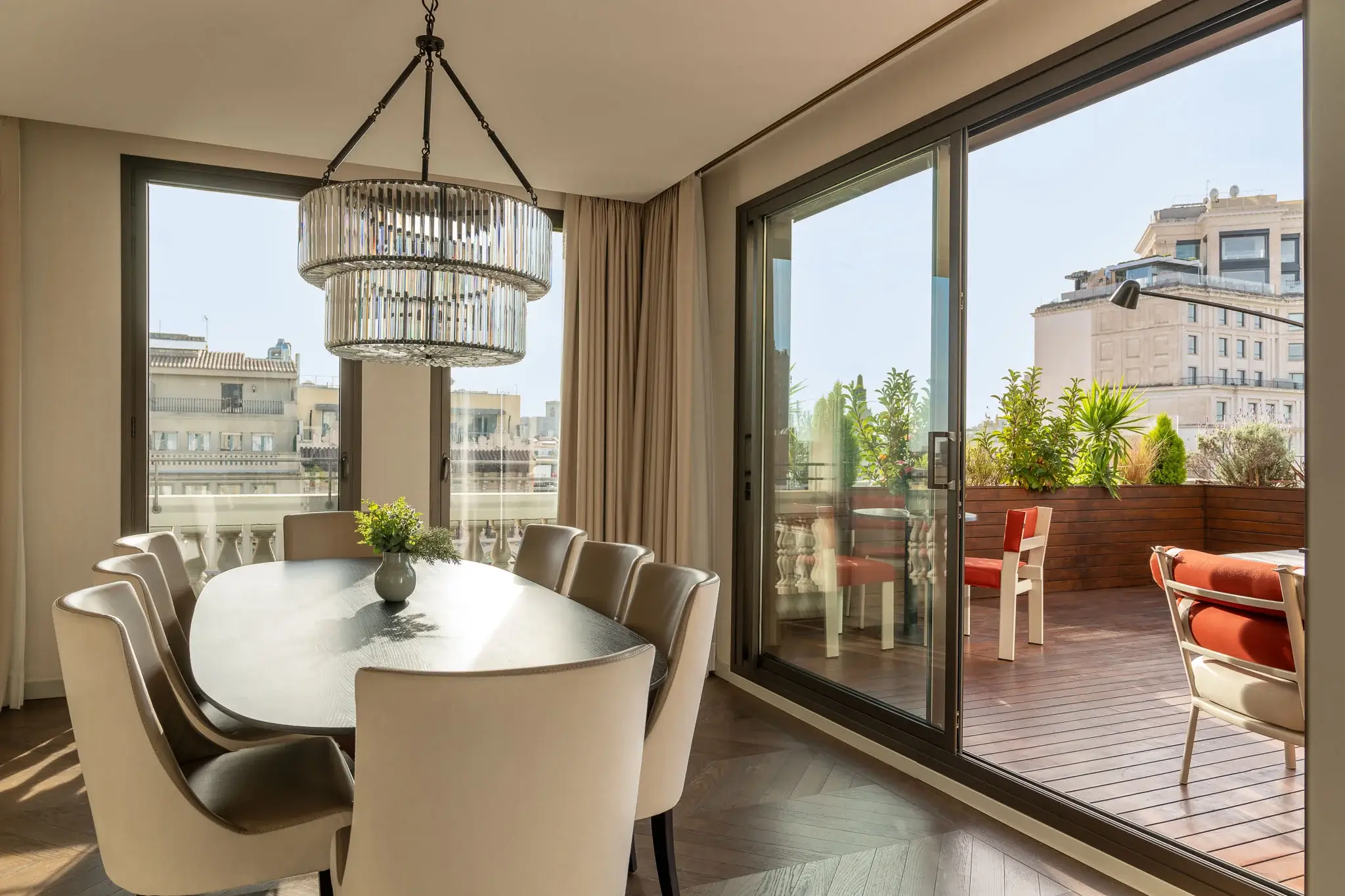 Modernes Esszimmer in einer luxuriösen Penthouse-Suite mit einem schwarzen ovalen Tisch, acht weißen Stühlen, einem runden Kronleuchter und großen Glastüren, die auf einen Balkon mit Pflanzen und Sitzgelegenheiten im Freien führen.