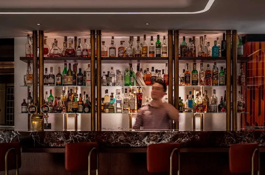 A bartender shakes a cocktail behind a marble bar, with shelves of assorted liquor bottles displayed in the background.