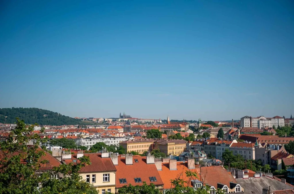 Cityscape view with red-roofed buildings, greenery, and distant hills under a clear blue sky.