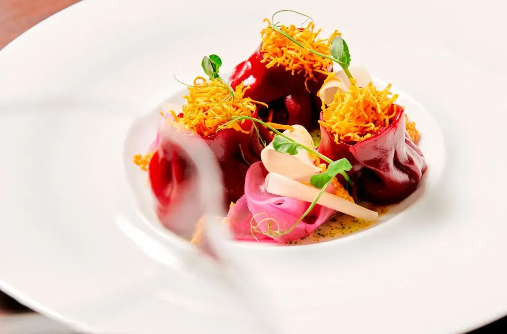 A plated gourmet dish perfect for meetings or events, featuring red pasta parcels, crispy yellow strips, fresh greens, and thinly sliced vegetables on a white plate.