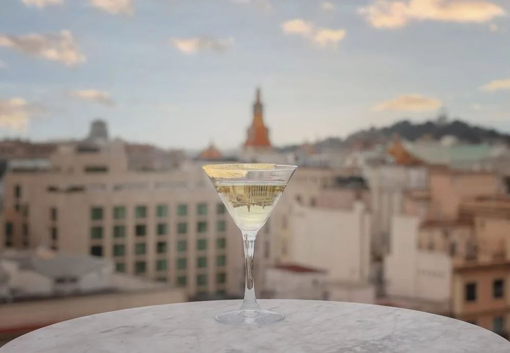 Una copa de martini plena d'una beguda clara es troba sobre una taula rodona de marbre a Terrassa Azimuth, amb un paisatge urbà borrós i un cel ennuvolat al fons.