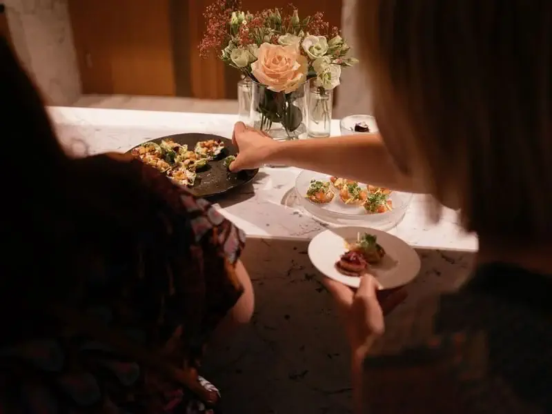 Dva lidé stojí u mramorového pultu s talířem předkrmů Externí Catering, jeden se natahuje po jídle, zatímco druhý drží talíř; v pozadí je vidět váza s květinami.