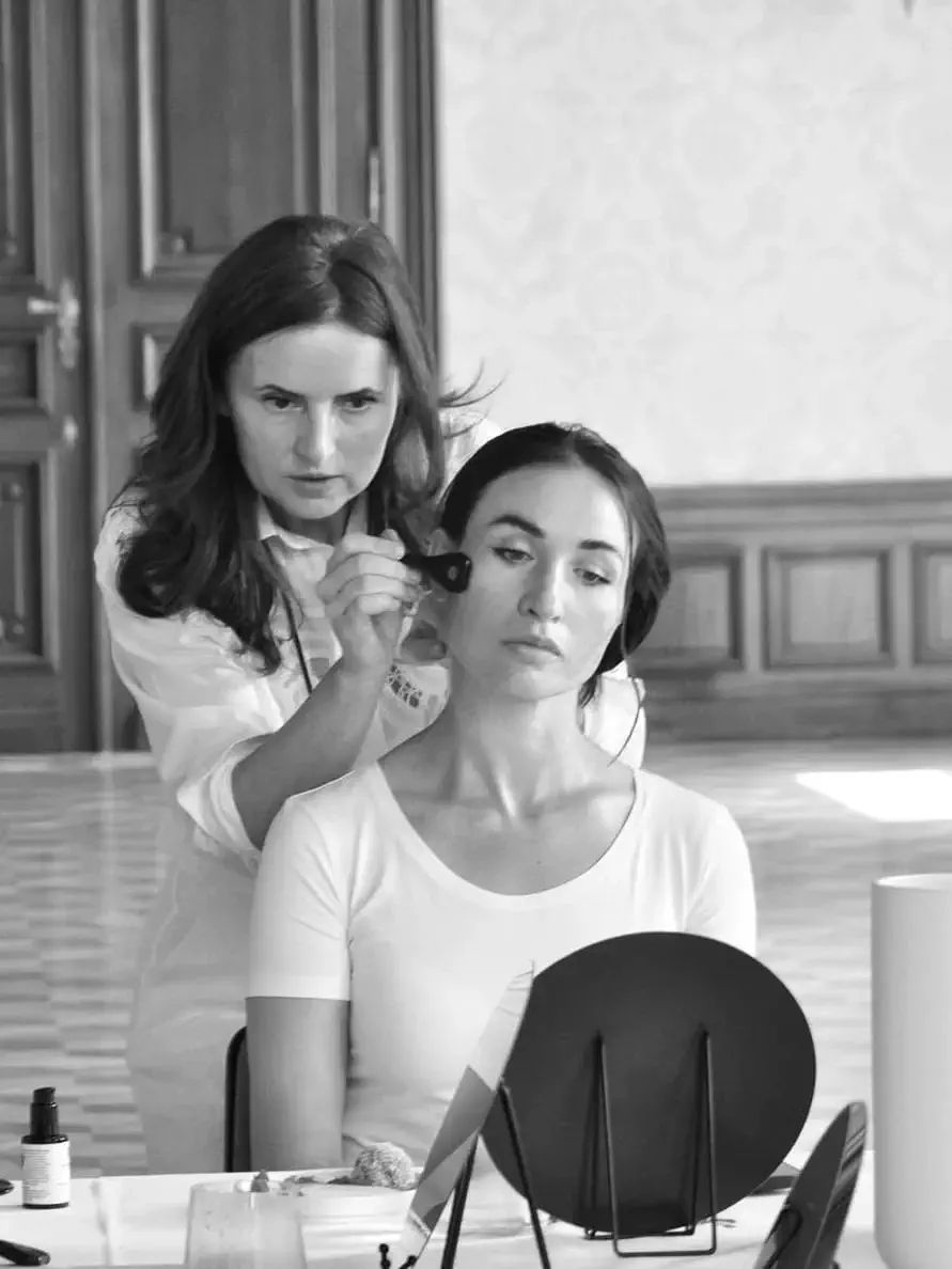 A woman from Me Myself Vienna applies makeup to another woman seated in front of a mirror in an elegant room with wooden paneling.