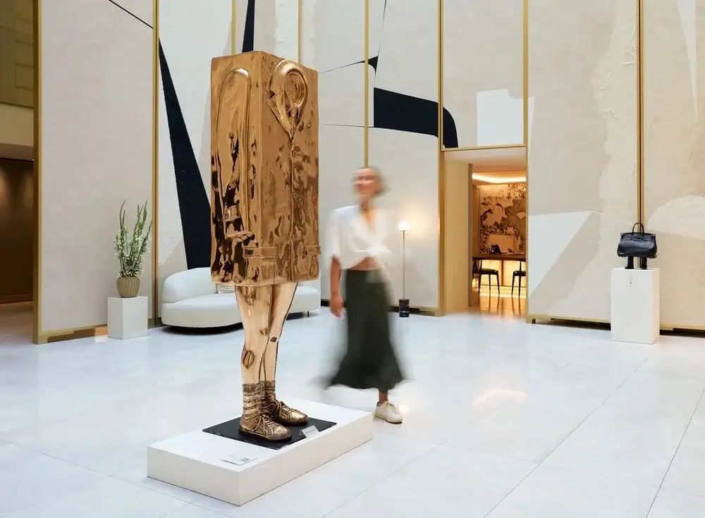 A person walks past a tall, gold-toned abstract sculpture featuring a coat and shoes in a modern, minimalist lobby.