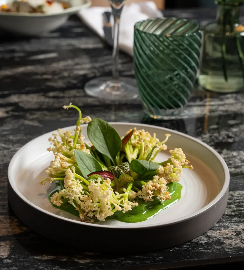 Un plato blanco del restaurante Virens, con salsa verde, hojas verdes y racimos de pequeñas flores blancas, está sobre una mesa de mármol oscuro junto a un vaso verde.