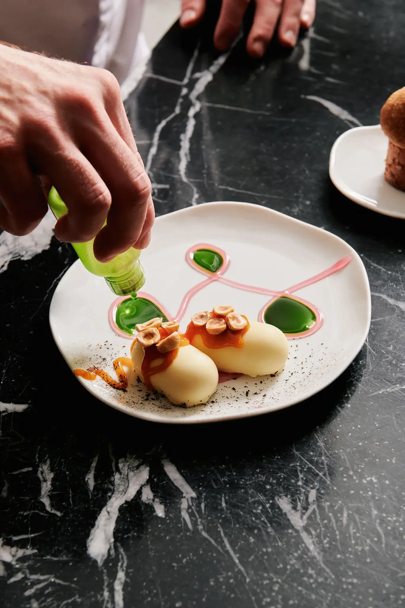 A person decorates a plated dessert with sauce using a squeeze bottle; the dish features two plant-forward, oval-shaped pastries with toppings on a white plate.