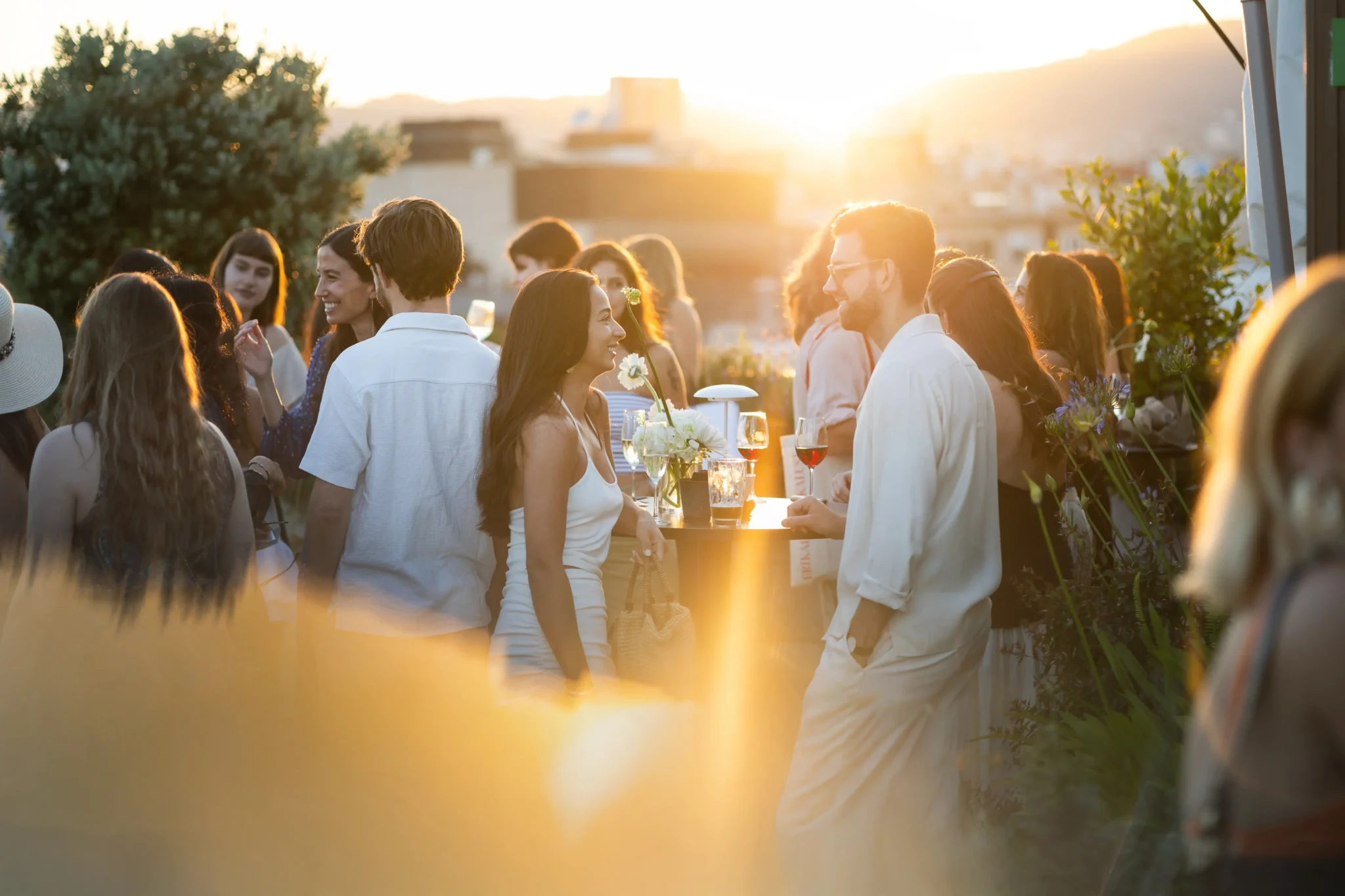 Un grupo de personas se reúne al aire libre al atardecer para asistir a eventos sociales, de pie y charlando alrededor de mesas con bebidas mientras la luz del sol crea una atmósfera cálida y resplandeciente.