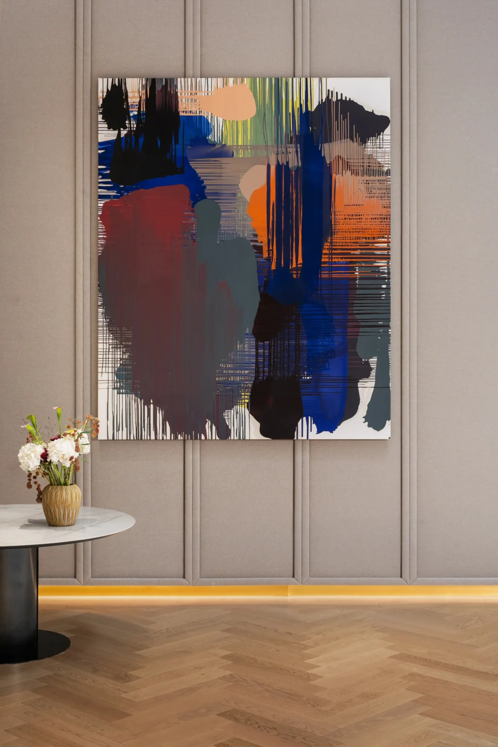 Large abstract painting with bold blue, red, black, and orange brushstrokes hangs on a beige paneled wall above a herringbone wood floor at Almanac Palais Vienna, with a round table and flower vase completing the elegant scene.