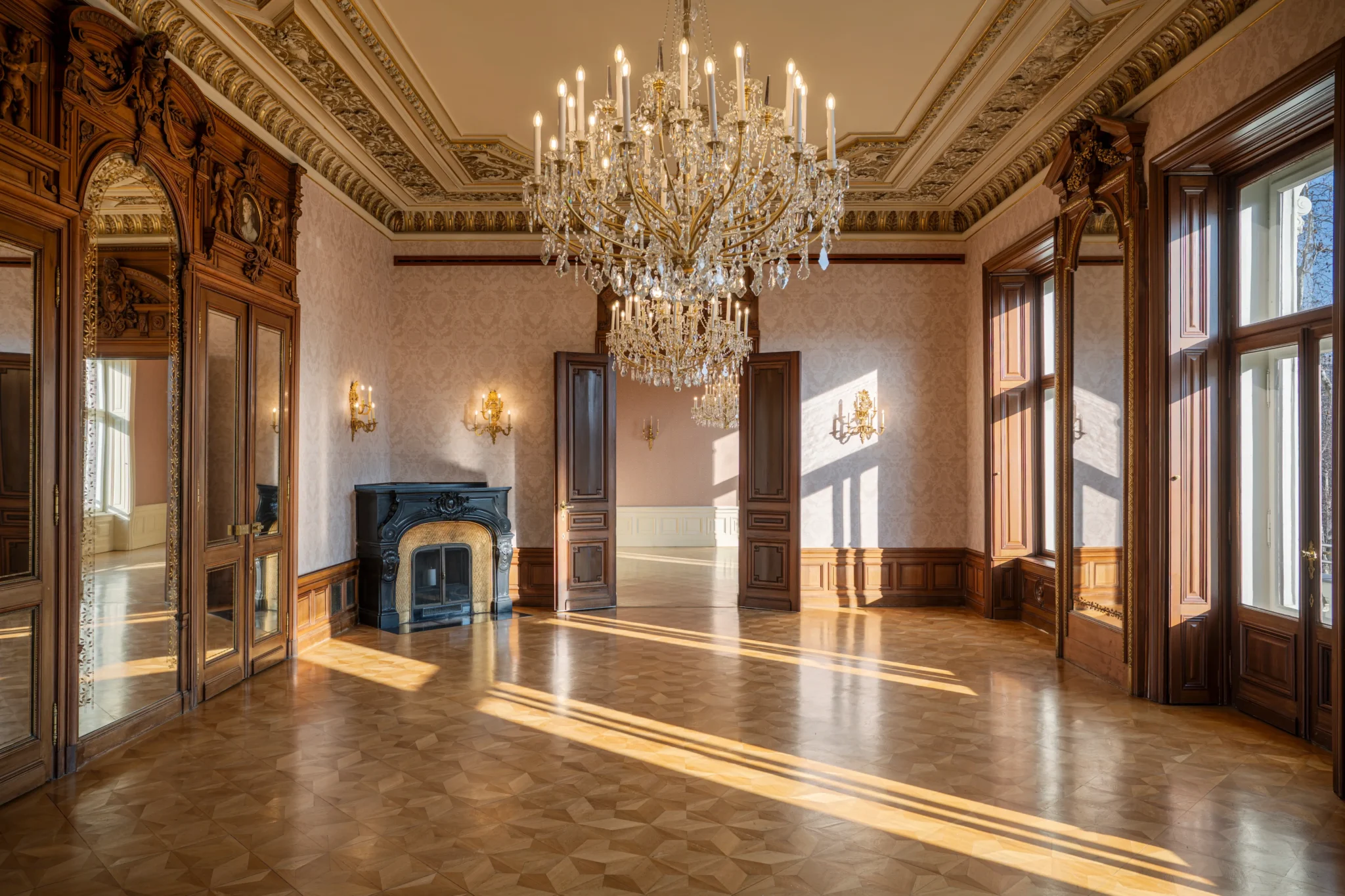 Elegant, empty ballroom with intricate woodwork, a black fireplace, large windows, ornate ceiling, and two crystal chandeliers hanging from the ceiling. Sunlight streams across the parquet floor.