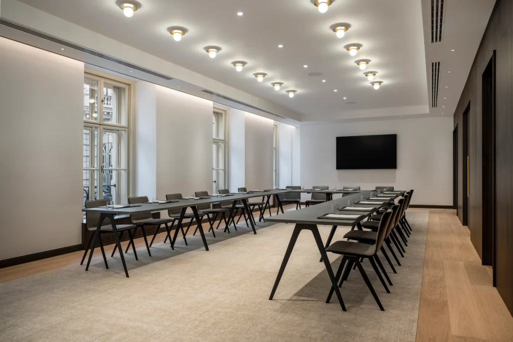 A modern conference room with two rows of tables and chairs, a large screen on the wall, notebooks and glasses set on tables, and large windows along one side.