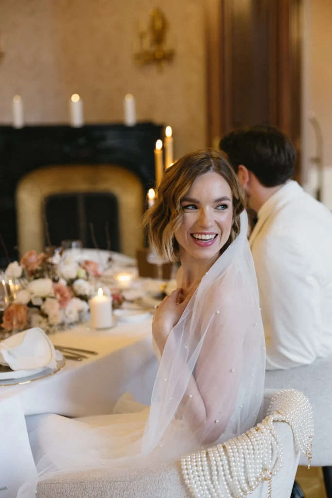 A bride in a white dress and veil smiles while seated at a decorated table with candles and flowers at a special event; a man in a white suit sits beside her.